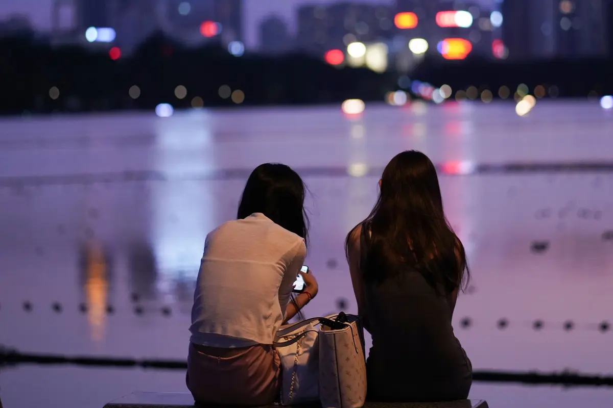 Dos mujeres sentadas de espaldas junto a un lago, mirando las luces de la ciudad al anochecer